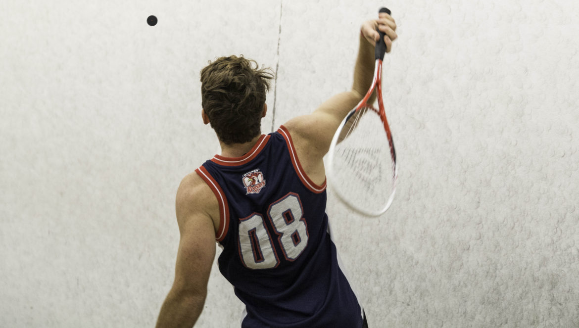 Male jumping to hit a squash ball with a squash racket. Person is wearing a sport singlet with 08 on the back.